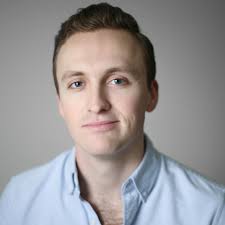Smiling man with short brown hair and blue eyes wearing a light blue shirt.